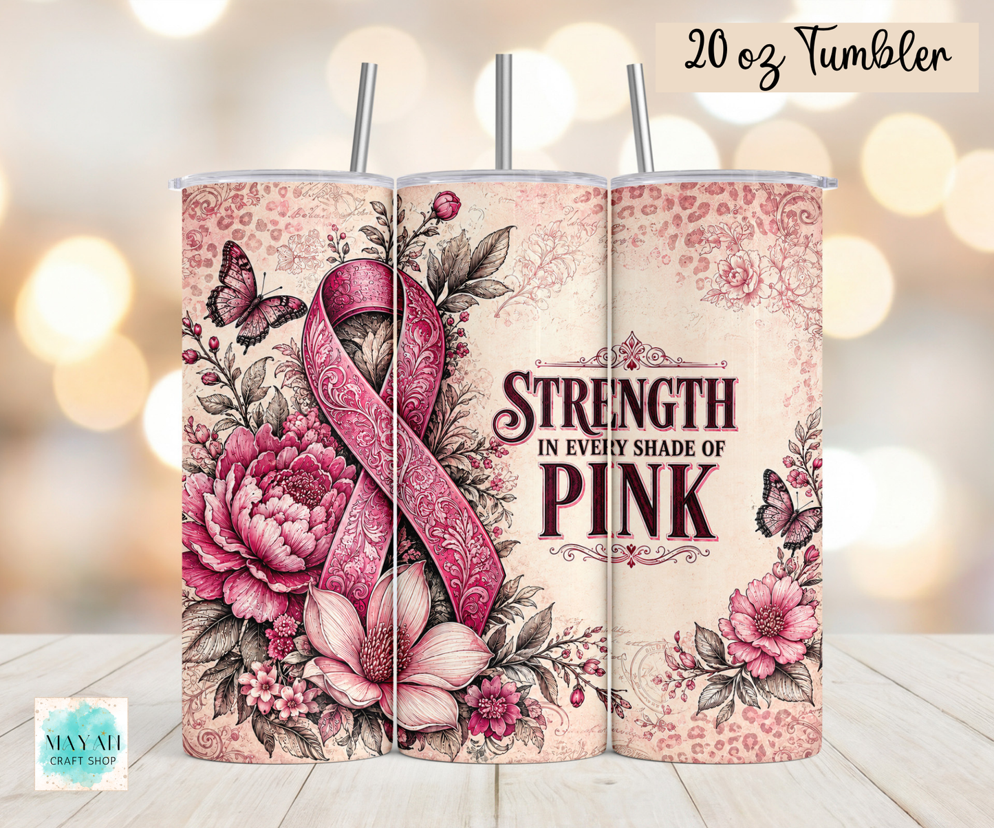 A 20 oz tumbler shown in three wrap-around views on a light wood surface with a blurred warm bokeh background. The design features a pink awareness ribbon, pink flowers, butterflies, and the text 'STRENGTH IN EVERY SHADE OF PINK.