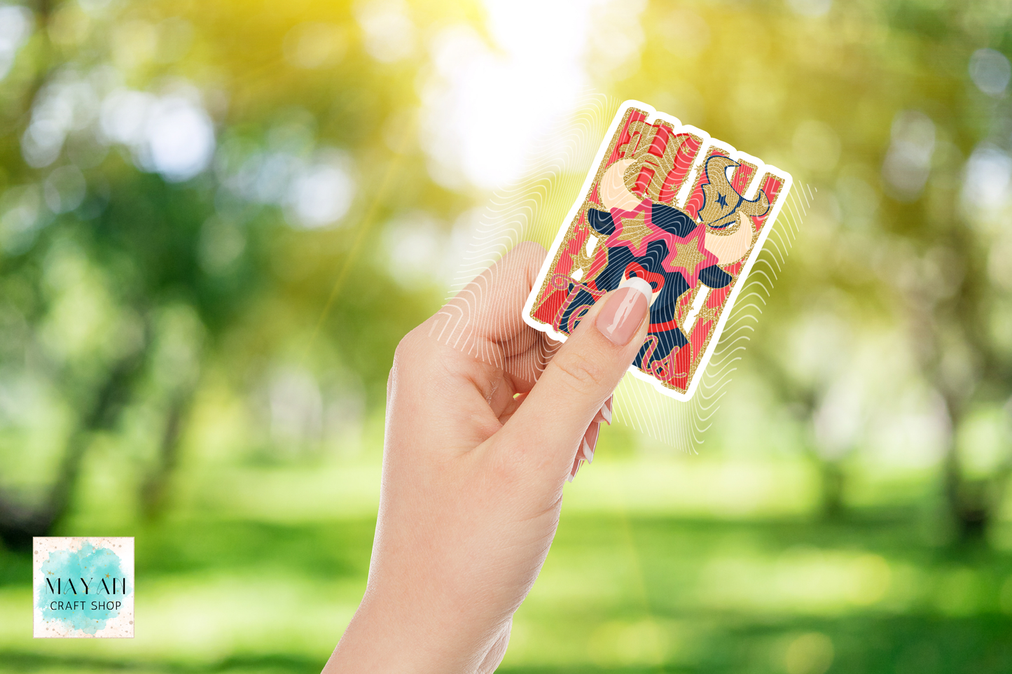 A hand holds a colorful sticker featuring a bear and a text against a bright, sunlit green park background.