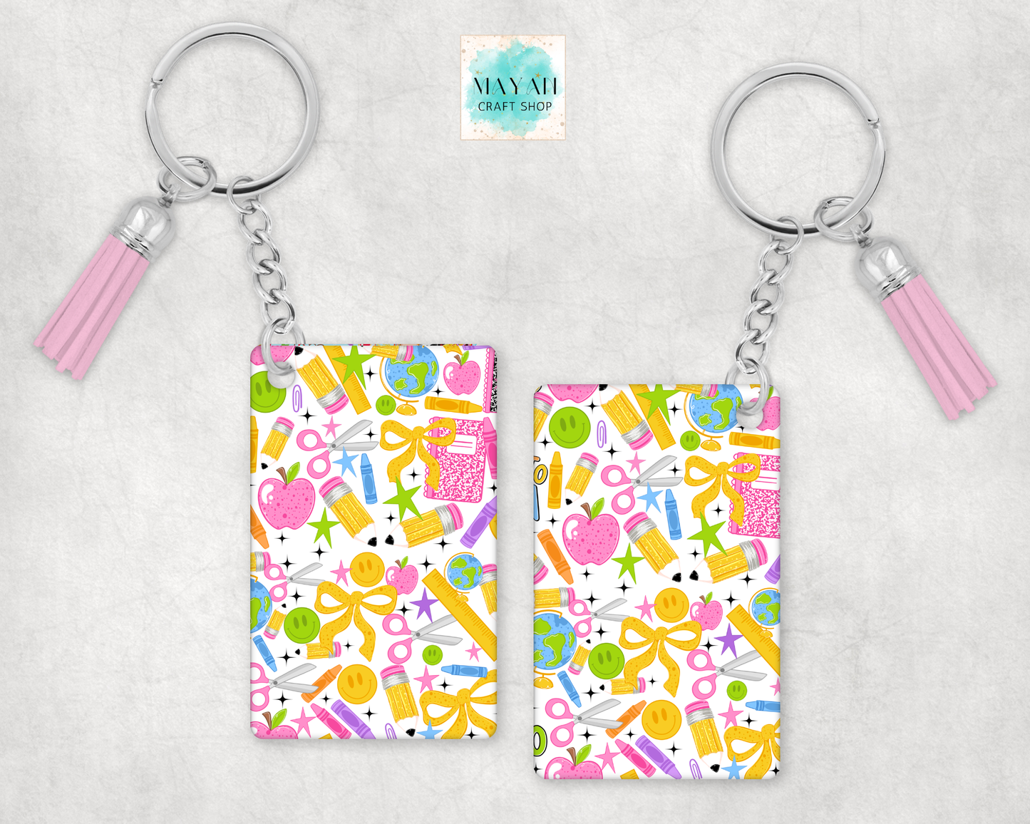 Two rectangular acrylic keychain charms with a colorful school supply pattern, attached to silver keyrings with pink tassels, displayed on a light gray textured background.