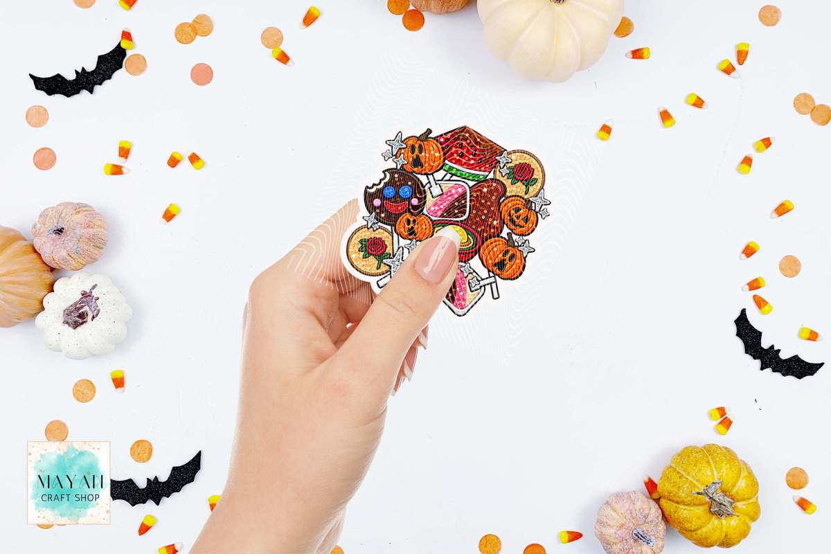 A hand holds a cluster of Halloween-themed stickers featuring pumpkins, ghosts, candy corn, and a witch hat, set against a white background decorated with small pumpkins, candy corn, and black bat cutouts.