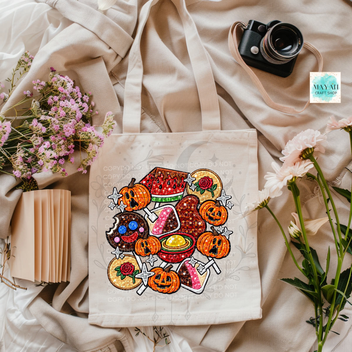 A beige canvas tote bag with a colorful Halloween-themed graphic print, styled in a flat-lay arrangement with a vintage camera, pink flowers, a small notebook, and draped cream fabric.