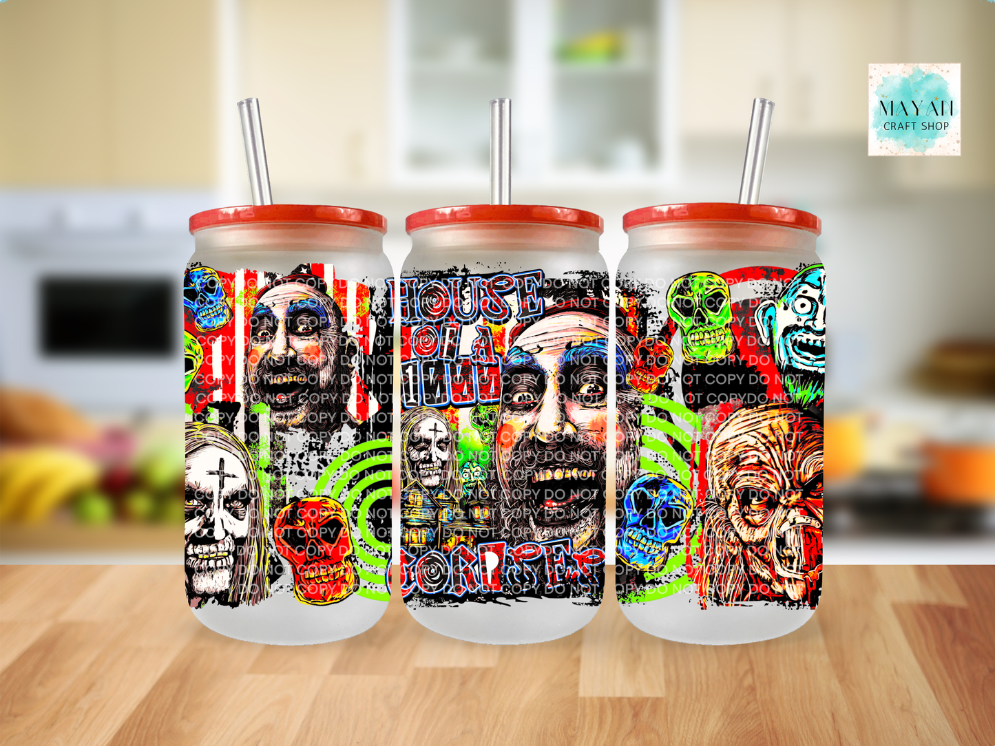 Three frosted glass tumblers with red lids and clear straws, featuring colorful horror-themed graphic prints, displayed on a wooden table with a blurred kitchen background.