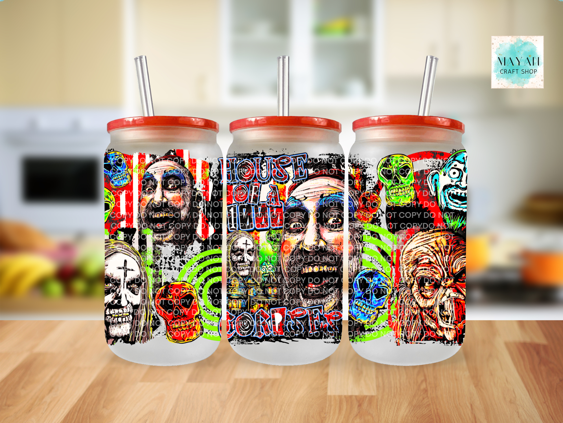 Three frosted glass tumblers with red lids and clear straws, featuring colorful horror-themed graphic prints, displayed on a wooden table with a blurred kitchen background.