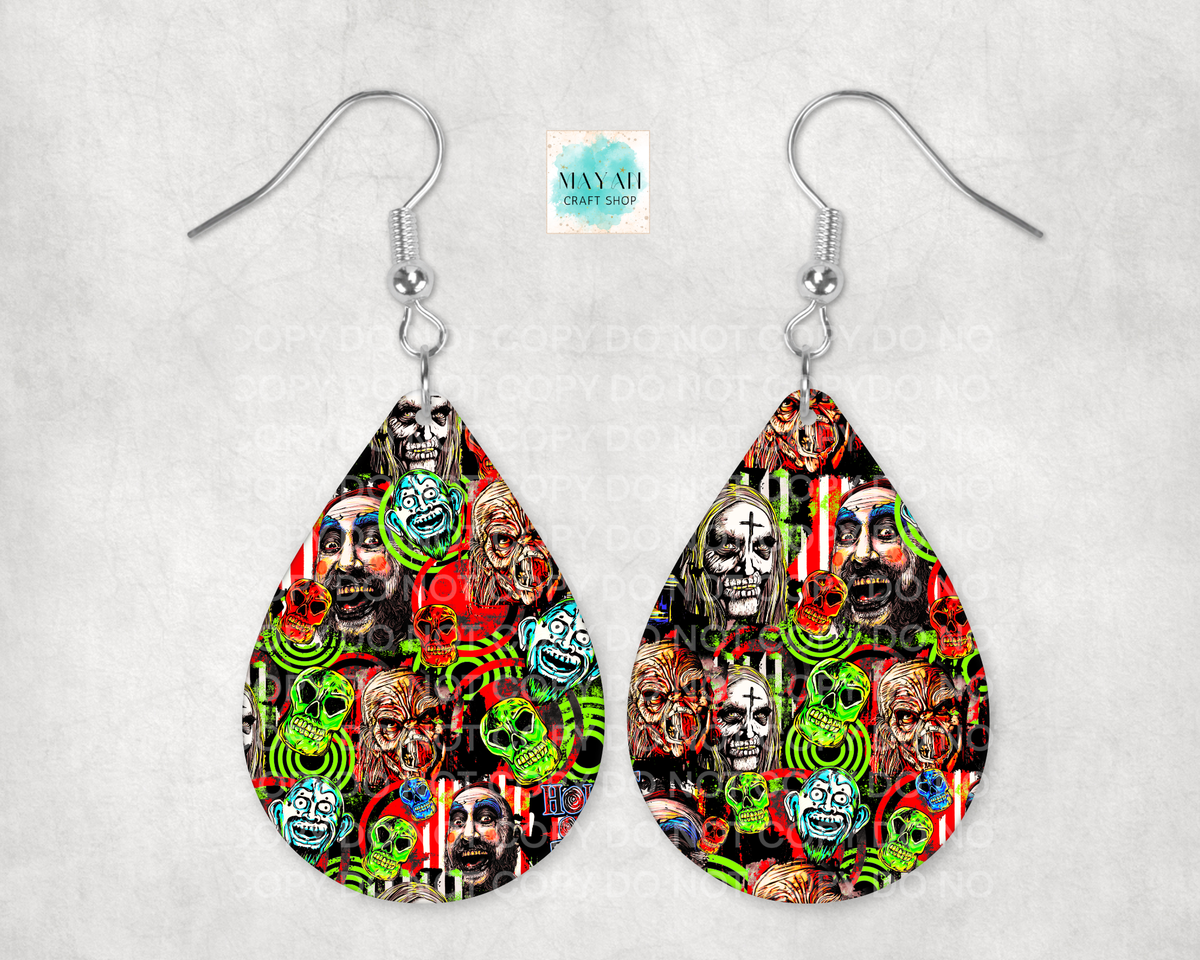 A pair of silver-tone dangle earrings with teardrop-shaped charms featuring a dense, colorful collage print of skulls, faces, and neon green spiral motifs on a black background, displayed against a light gray textured backdrop.
