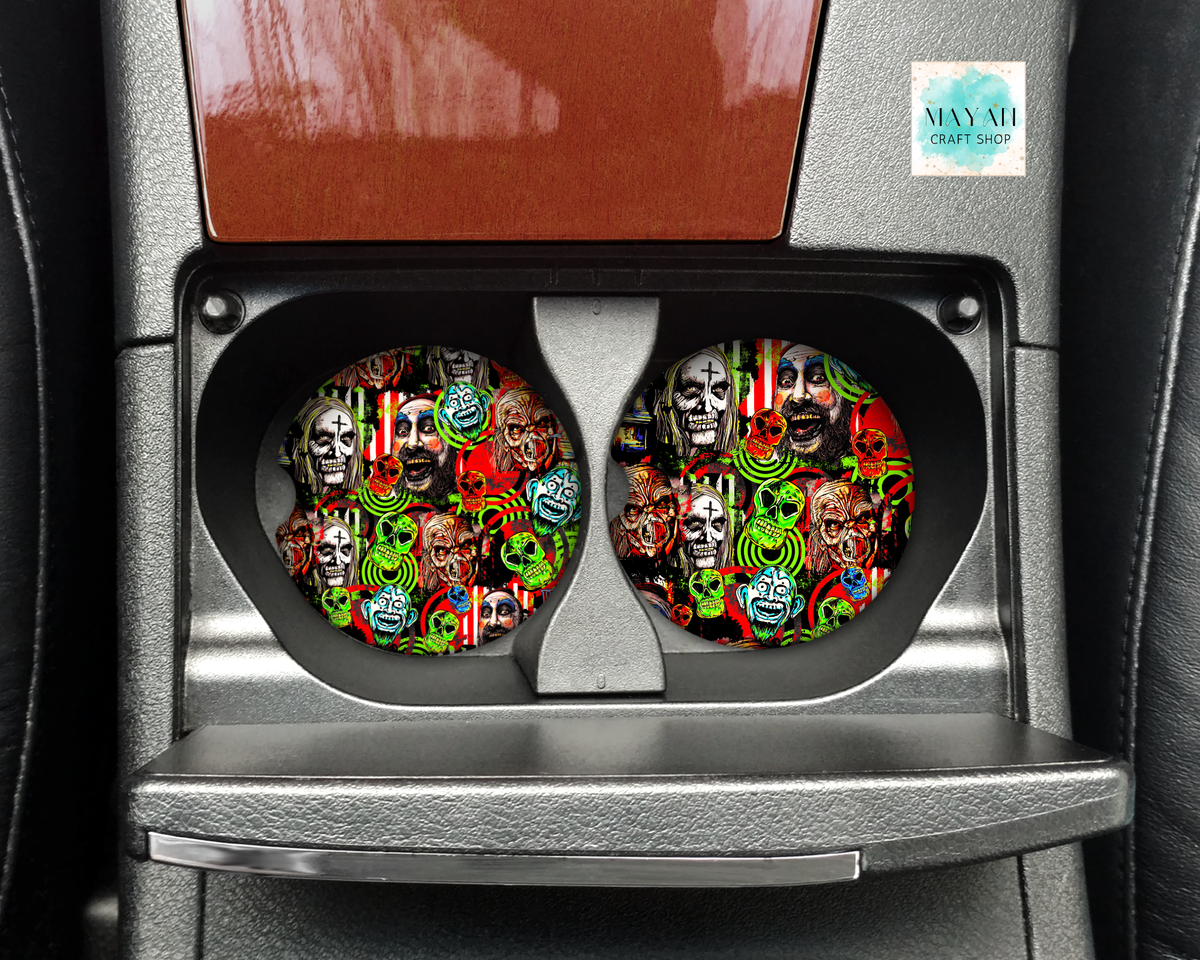 Two circular car cup holders installed in a center console, featuring a colorful, busy collage pattern of skulls, masks, and psychedelic designs. The cup holders are set inside a dark gray textured car interior.