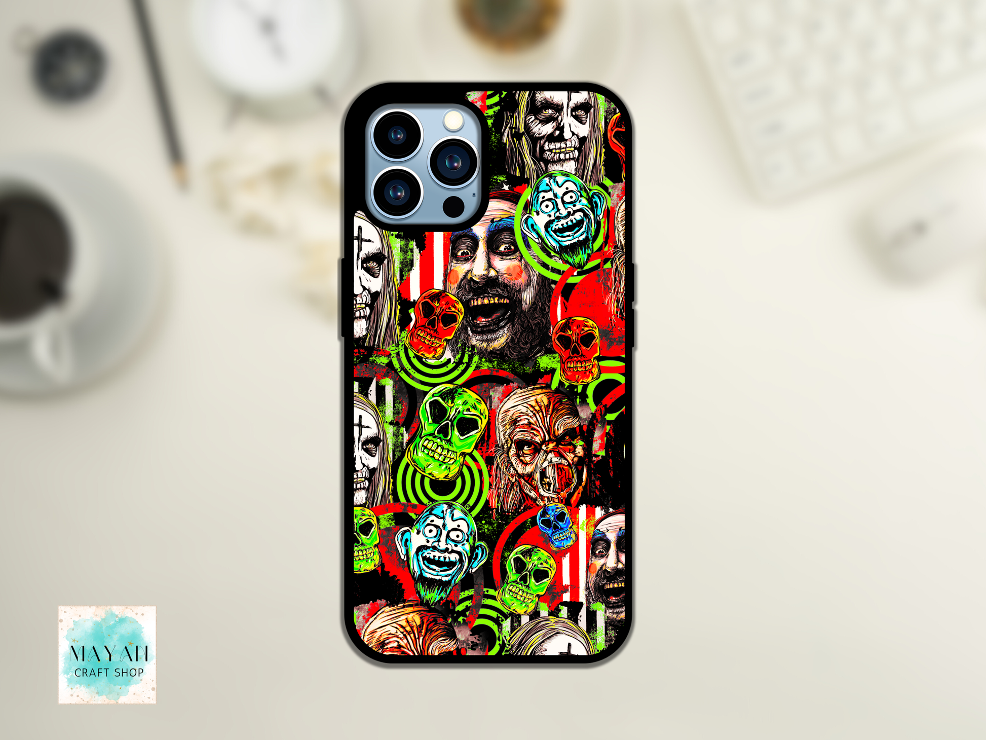 A phone case featuring a highly detailed, colorful collage of horror clown and zombie faces, displayed on a light-colored desk with a blurred keyboard and coffee cup in the background.