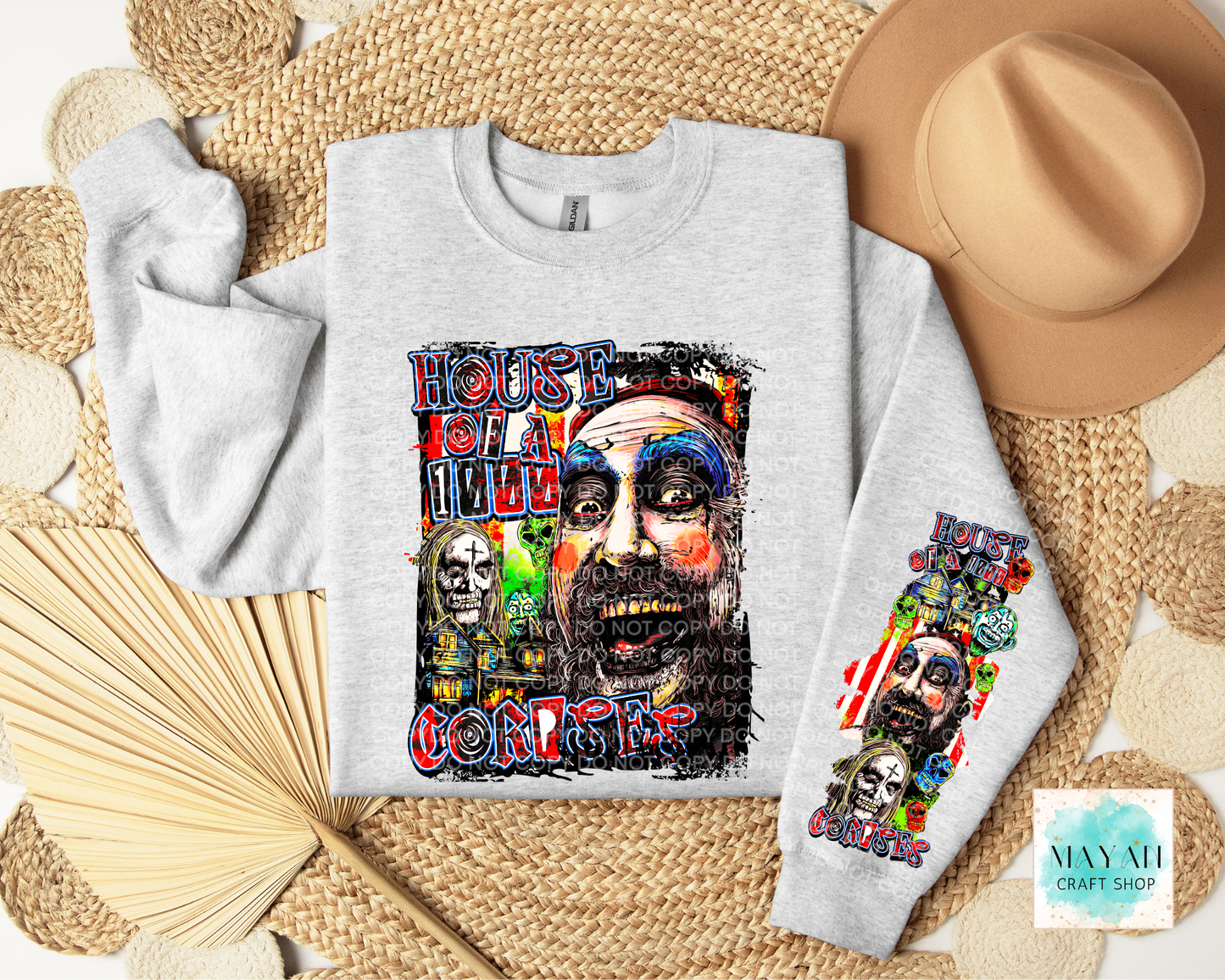 A light heather gray crewneck sweatshirt laid flat on a woven mat, featuring a large colorful graphic print on the front and a smaller matching graphic on the sleeve. A tan hat, folded beige towel, and wooden fan surround the sweatshirt.