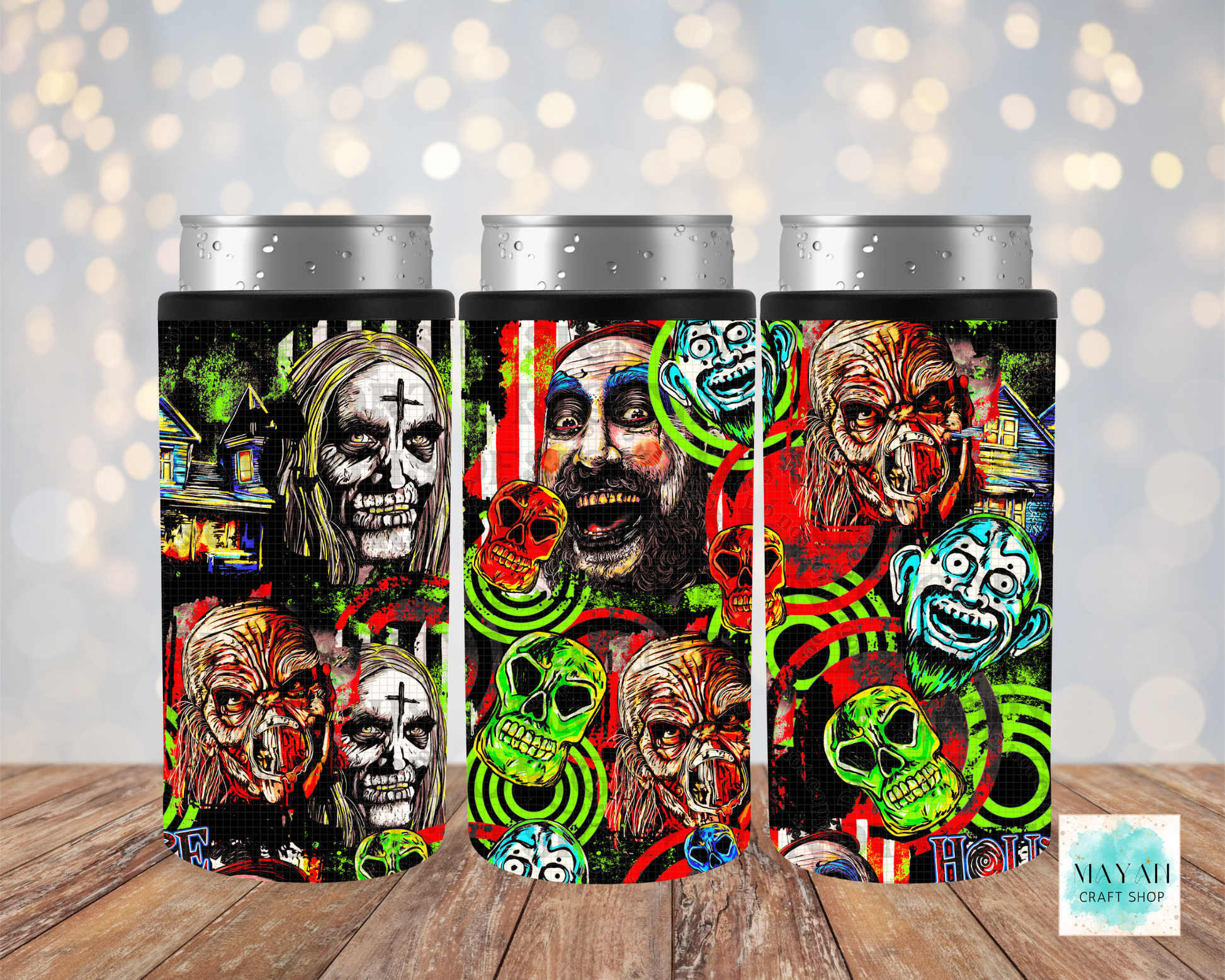 Three slim can coolers with silver aluminum can tops, featuring a vibrant, multi-colored Halloween collage print of skulls, clowns, and ghosts on a black background, displayed on a wooden surface with a soft bokeh light background.
