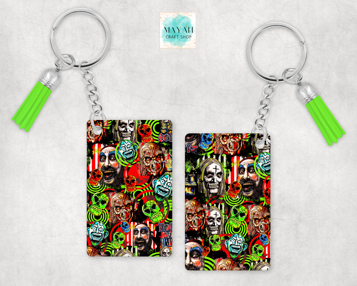 Two keychains with silver keyrings and bright green tassels, shown on a light gray textured background. The keychains feature a colorful collage print with skulls, peace signs, and faces.