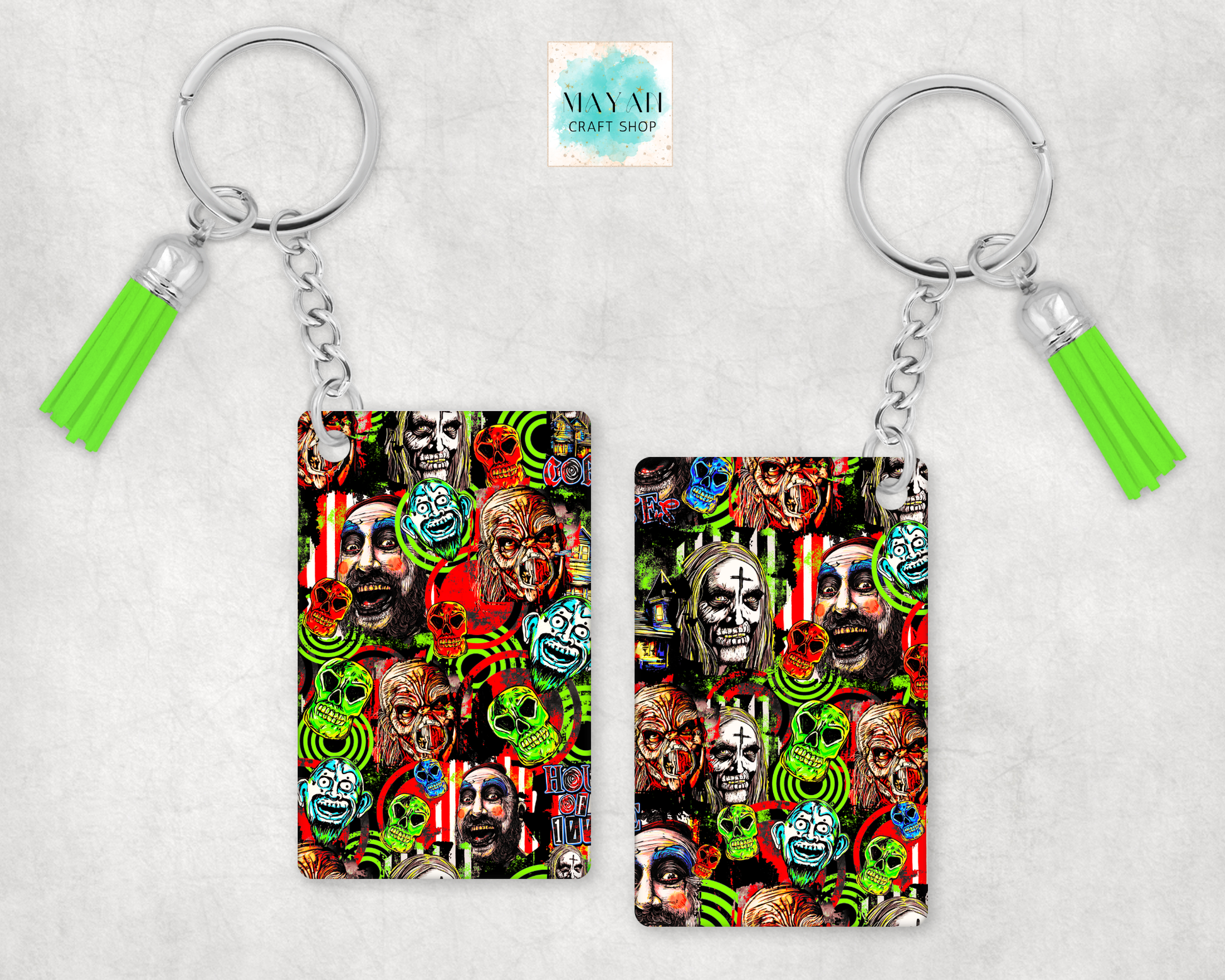 Two keychains with silver keyrings and bright green tassels, shown on a light gray textured background. The keychains feature a colorful collage print with skulls, peace signs, and faces.