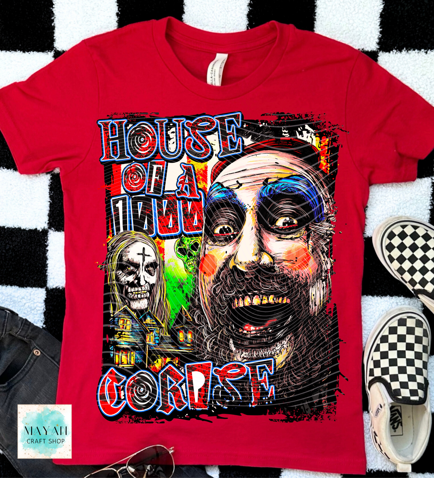 A red short-sleeve graphic T-shirt laid flat on a black-and-white checkered rug, with jeans, sunglasses, and checkerboard sneakers partially visible around it. The shirt features a large horror-style multicolor print and illustrated faces/skulls.