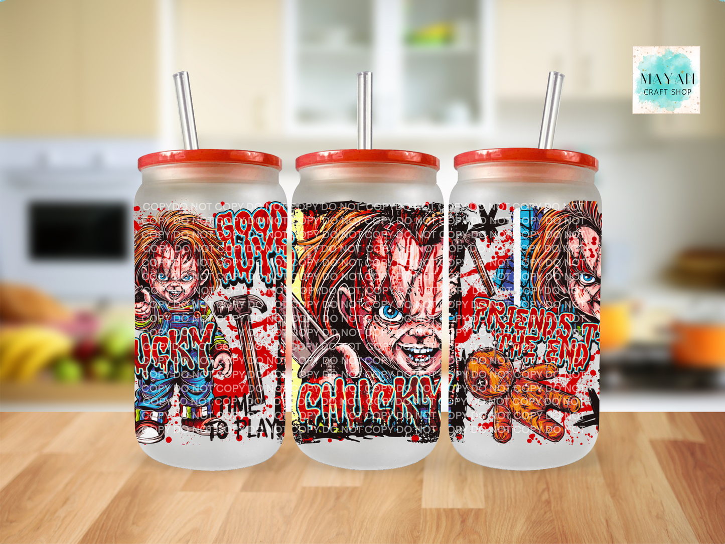 Three frosted glass tumblers with red lids and clear straws, featuring horror-themed artwork, displayed on a wooden surface with a blurred kitchen background.