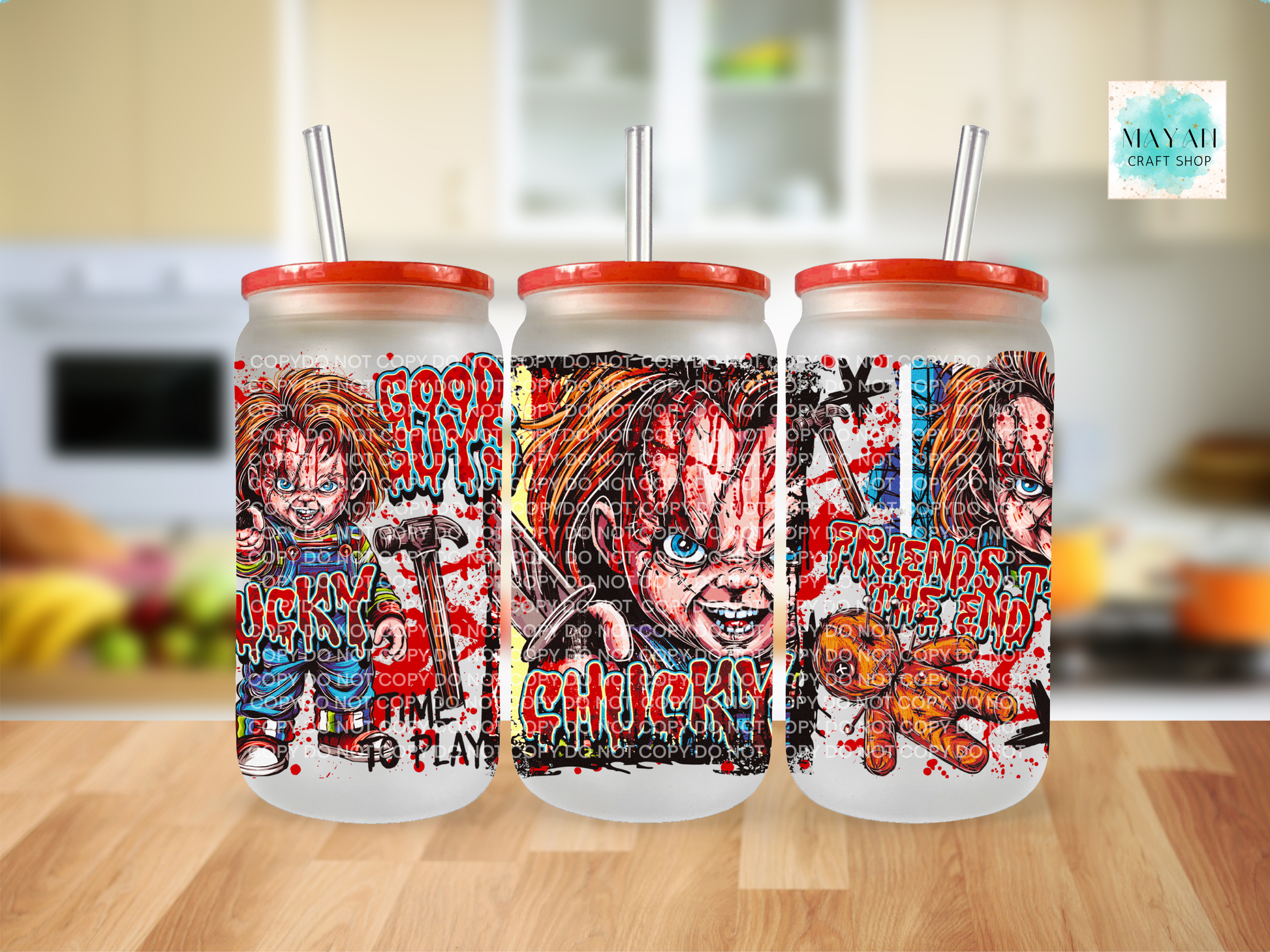 Three frosted glass tumblers with red lids and clear straws, featuring horror-themed artwork, displayed on a wooden surface with a blurred kitchen background.