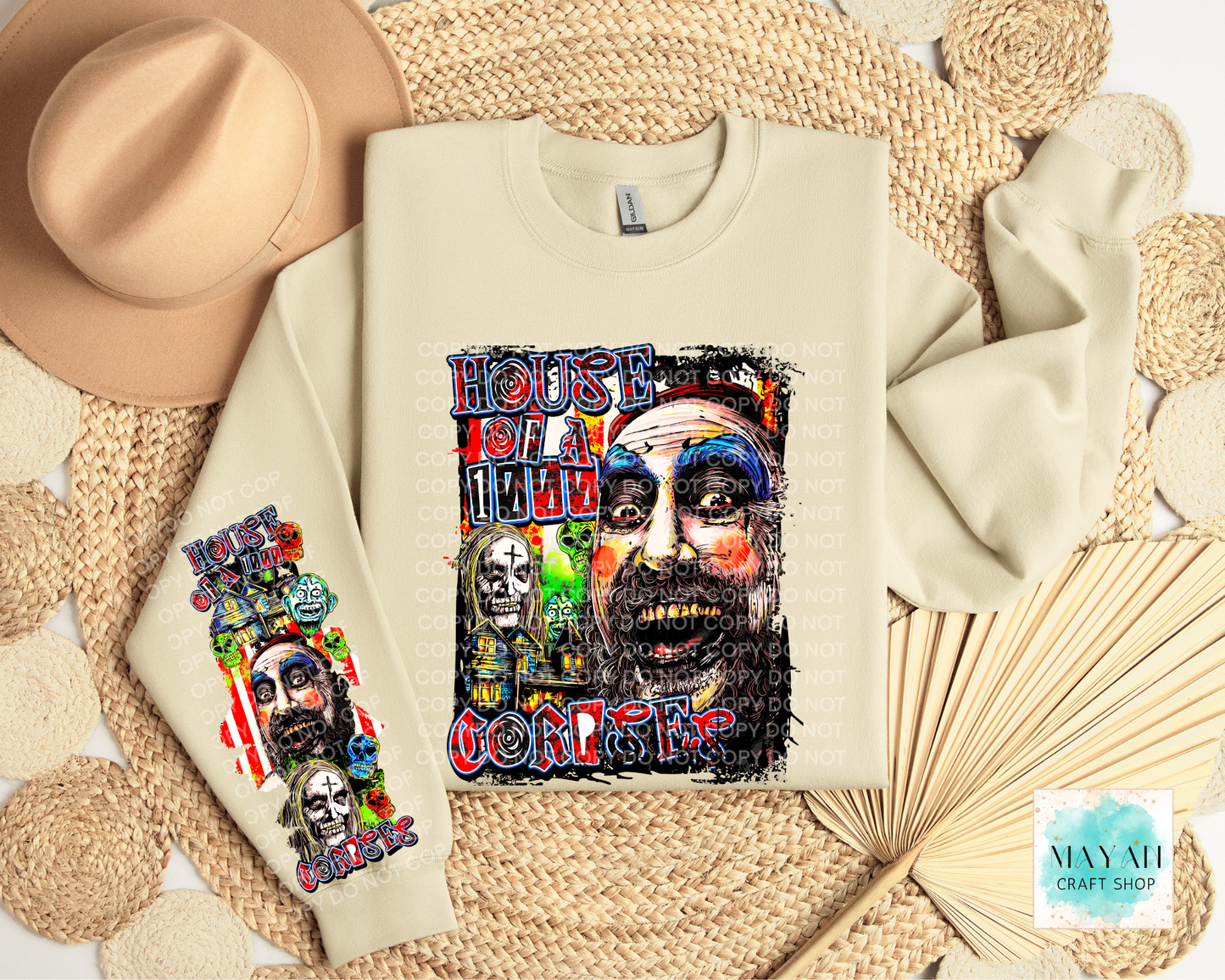 A cream-colored crewneck sweatshirt with a colorful graphic print on the chest, and a smaller matching graphic on the left sleeve. The sweatshirt is displayed in a styled flat-lay arrangement with a tan hat, woven placemats, and a paper fan.
