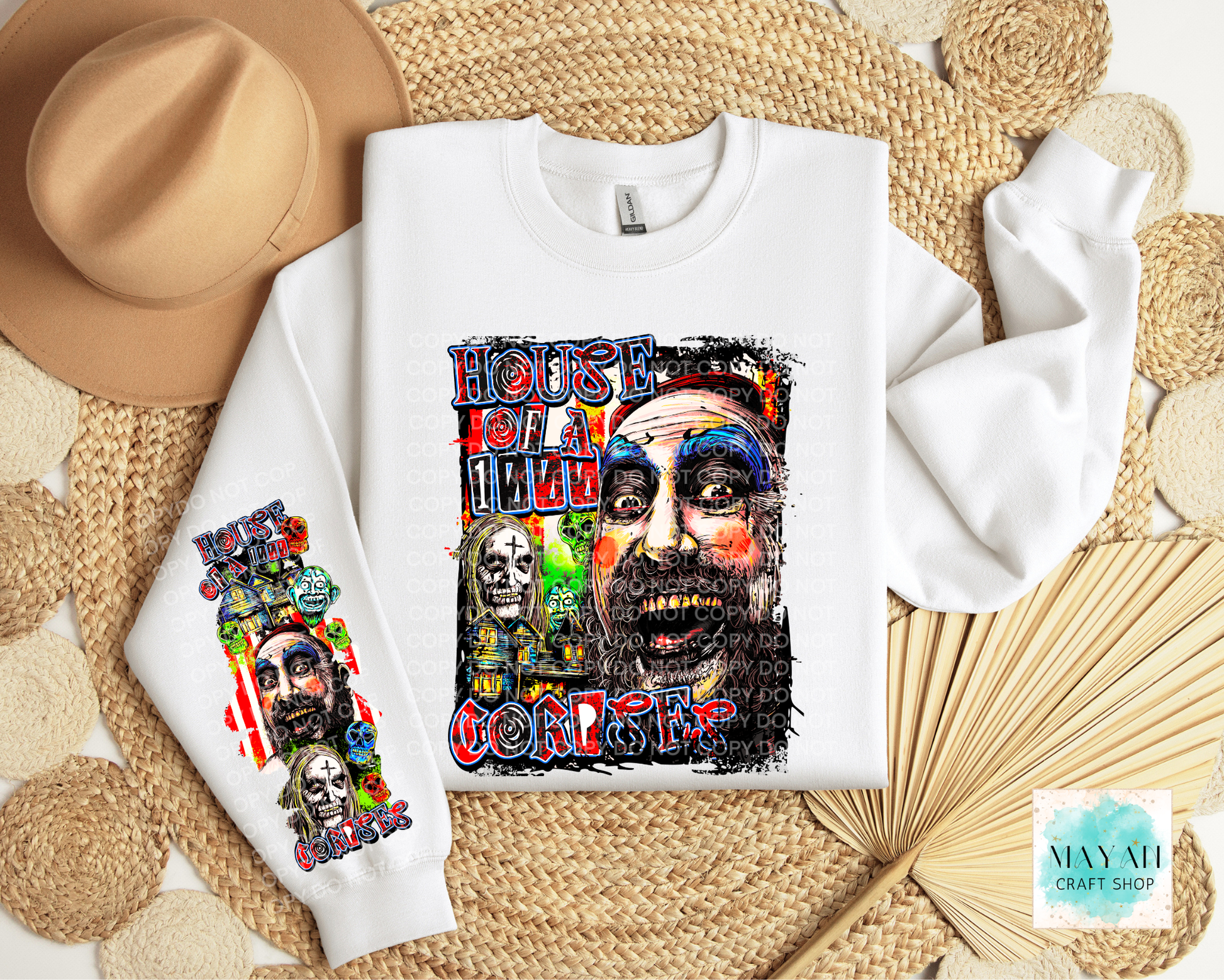 A white crewneck sweatshirt laid flat on a woven straw mat, featuring a colorful graphic print of a man's face on the front, with a matching sleeve print. A tan wide-brim hat and a folded cream towel are styled around it.