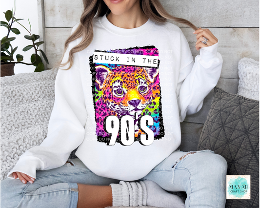 A woman wearing a white crewneck sweatshirt with a colorful graphic of a leopard face and the text 'STUCK IN THE 90'S', paired with light-wash ripped jeans, sitting on a gray couch.