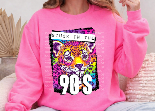 A person wearing a bright pink sweatshirt with a colorful graphic of a leopard face and the text 'STUCK IN THE 90's' on the front, seated indoors on a couch.