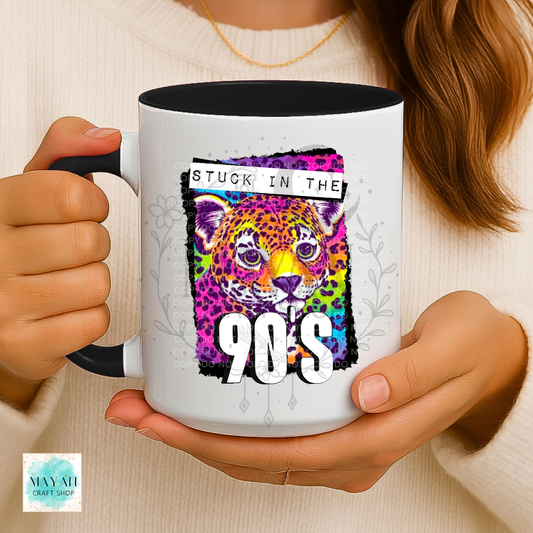 A person holds a white ceramic mug with black handles and interior. The mug features a colorful graphic of a leopard face with the text 'STUCK IN THE 90S' in large white letters.