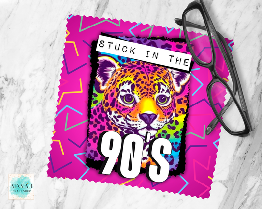 A lens cloth cleaner with a colorful retro design, featuring a rainbow leopard face with the text 'STUCK IN THE 90's' on a pink zigzag background. A pair of black eyeglasses rests on the top right corner of the napkin, which is laid flat on a white marble surface.