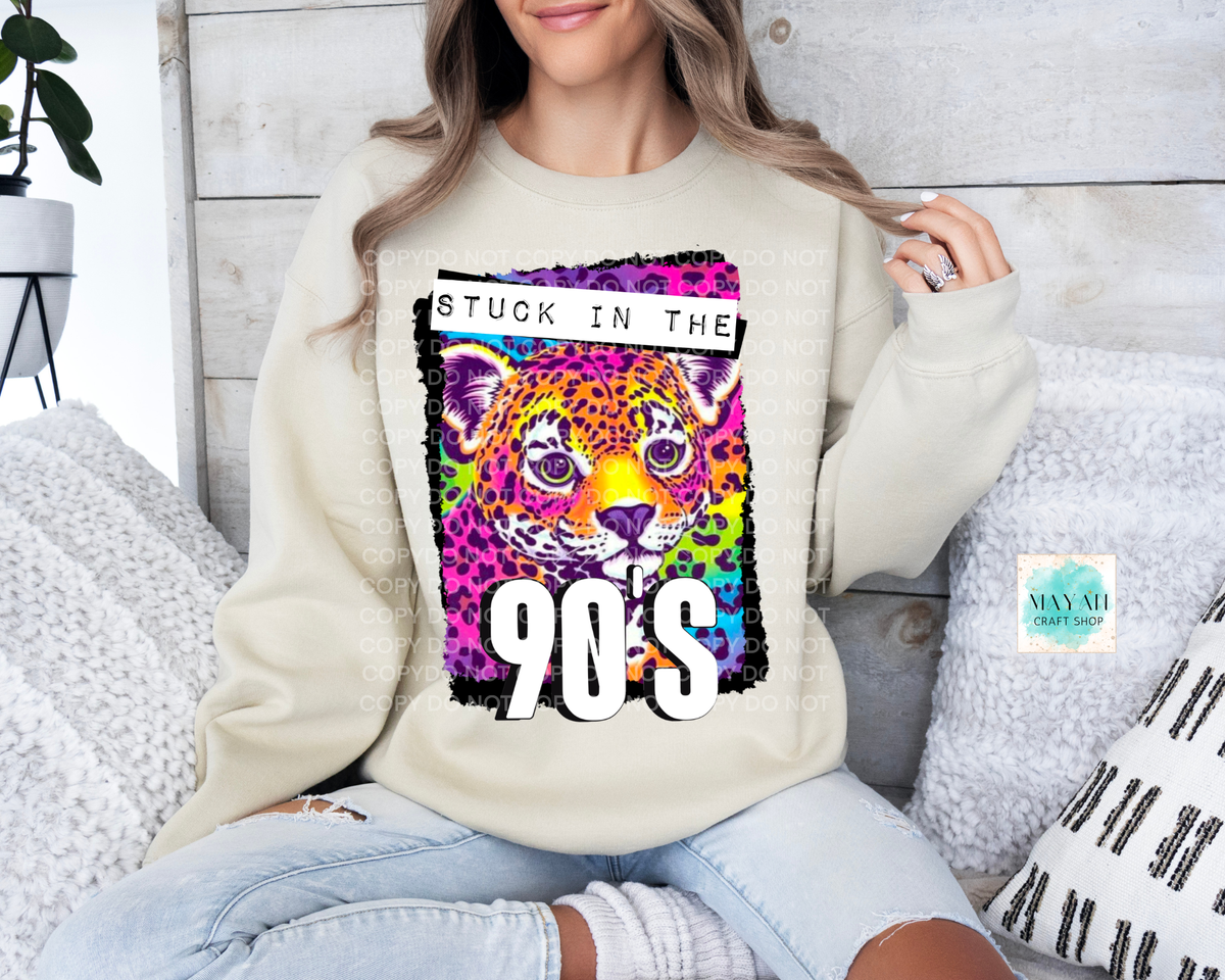 A woman wearing a cream-colored sweatshirt with a colorful graphic of a leopard and the text 'STUCK IN THE 90'S', paired with light-wash ripped jeans, sitting on a couch.