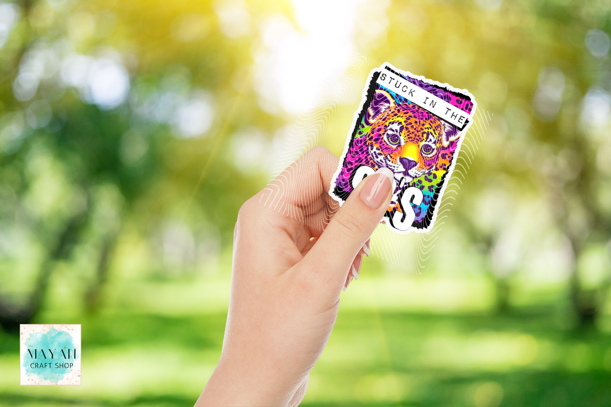 A hand holds a colorful die-cut sticker featuring a stylized tiger illustration and the text 'STUCK IN THE 90S' against a blurred green outdoor background.