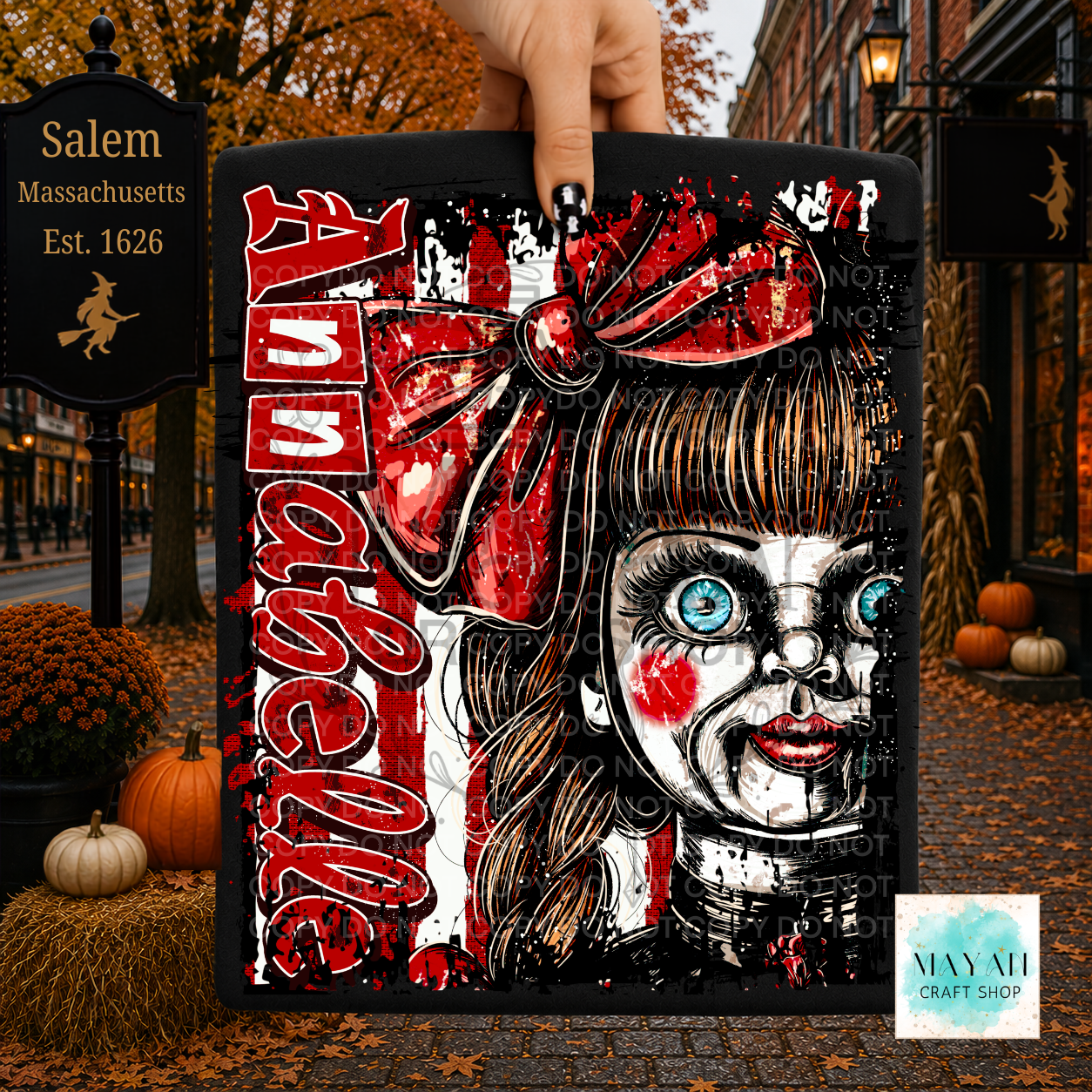 A hand holds a black square canvas featuring a stylized horror portrait of a doll-like girl with blue eyes, red bow. The background is a Halloween street scene with pumpkins, autumn leaves, and storefronts.