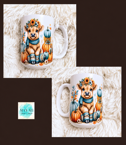 A white ceramic mug featuring a cute cartoon cow wearing a floral crown and blue scarf, surrounded by pumpkins and blue gourds. The mug is displayed in two views on a white faux fur background.