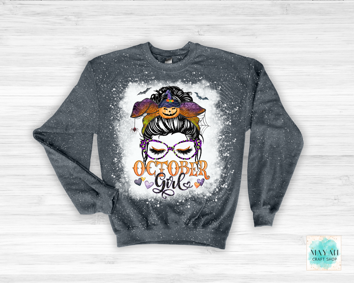 A dark gray heather crewneck sweatshirt with a distressed splatter finish, featuring a graphic print of a woman's face with glasses, a witch hat, pumpkins, and the text 'October Girl' in orange. The sweatshirt is laid flat on a white wood background.