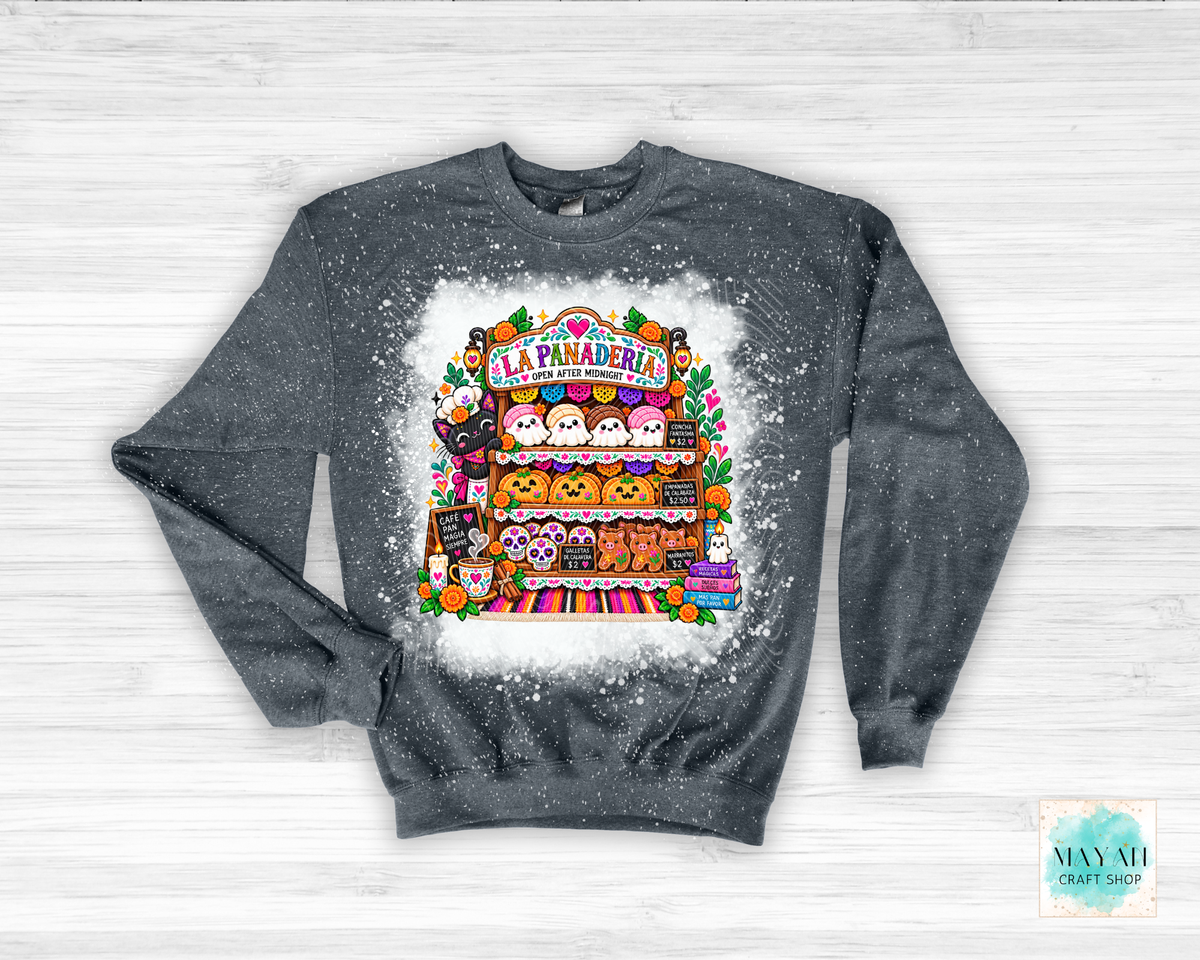 A dark gray heather crewneck sweatshirt featuring a colorful graphic of a Halloween-themed pastry shop named 'La Paraderia' with cute cartoon characters, displayed on a white wood background.