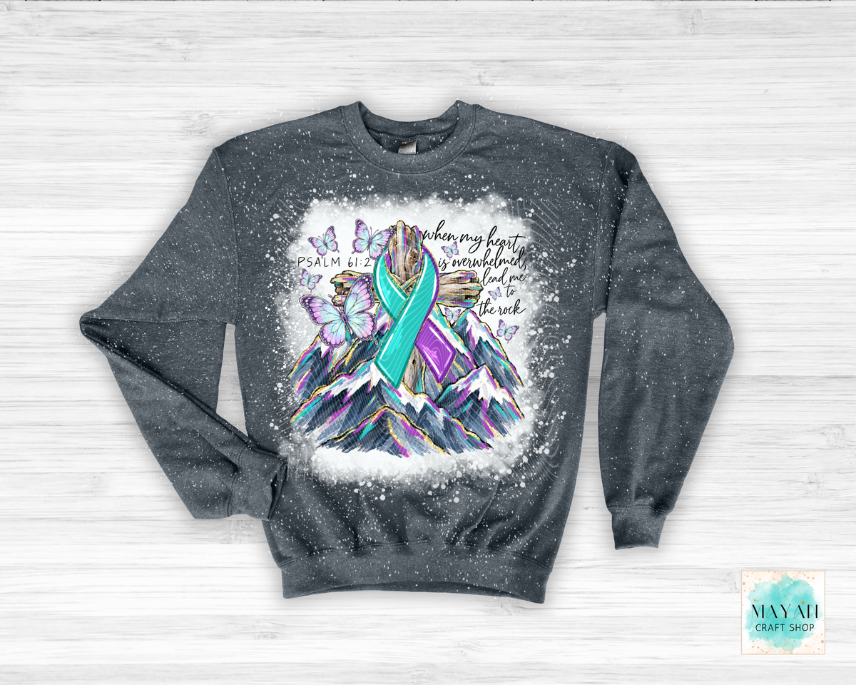A dark gray crewneck sweatshirt with a distressed splatter finish, featuring a colorful graphic of a mountain with a teal and purple ribbon, butterflies, and inspirational text. The sweatshirt is laid flat on a white wood background.