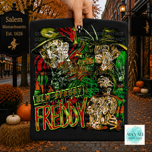 A hand holds a black shirt featuring stylized horror character artwork. The sign is photographed outdoors on a cobblestone street with autumn leaves, pumpkins, and storefronts in the background.