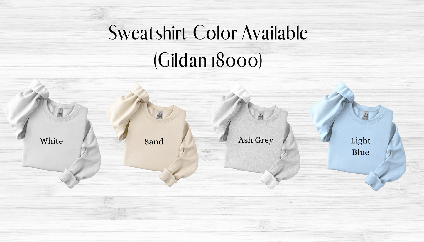 Four sweatshirts in white, sand, ash grey, and light blue on a wooden surface with text indicating color options.
