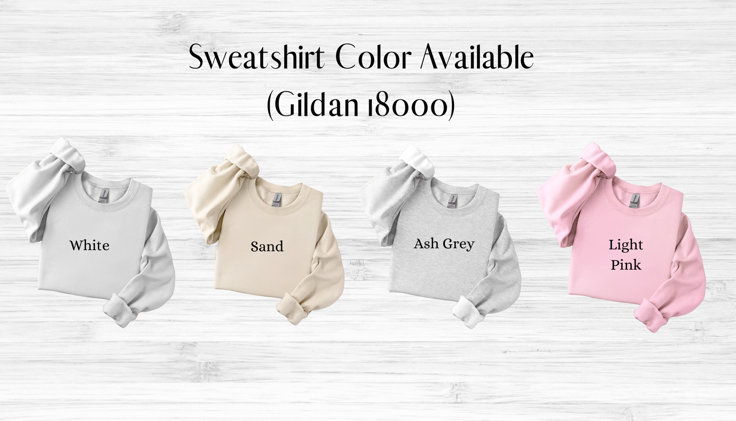 Four sweatshirts in white, sand, ash grey, and light pink on a wooden surface with text indicating color options.