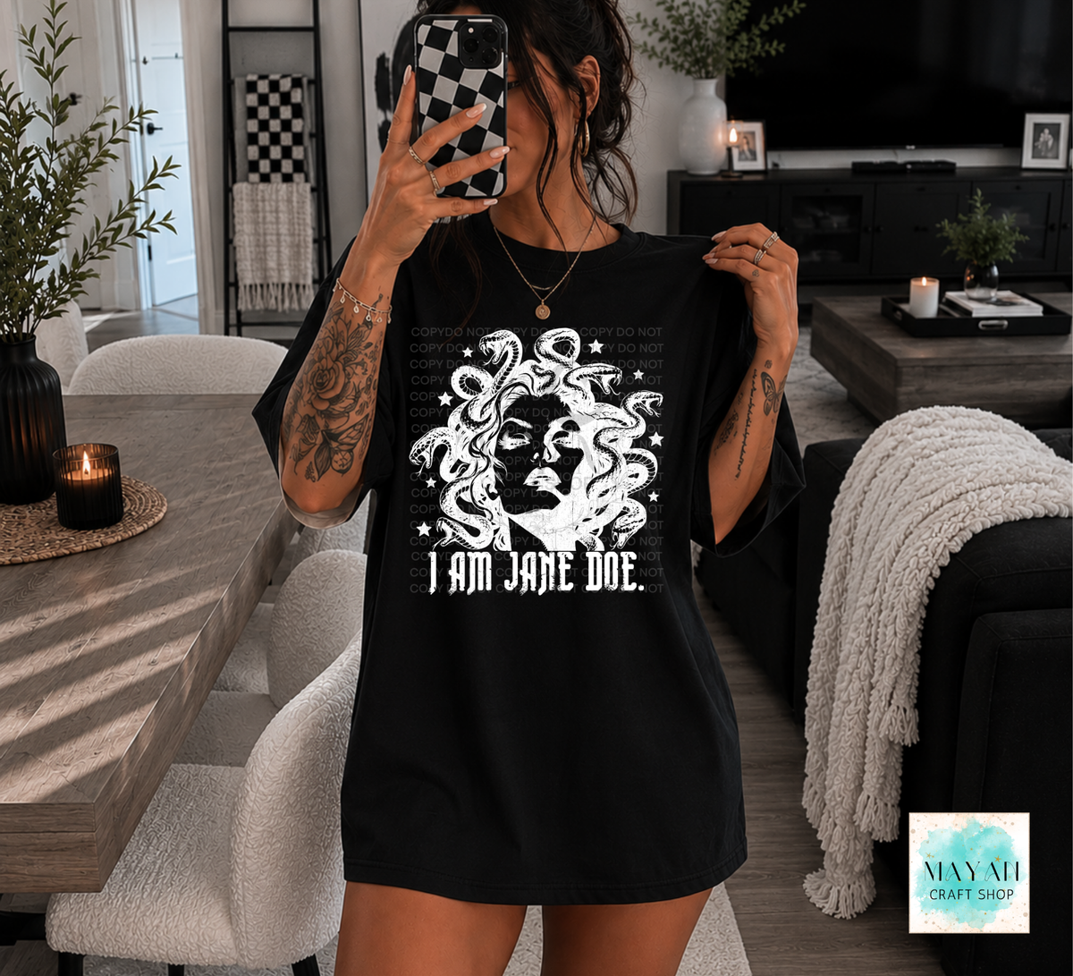 A woman takes a mirror selfie wearing a black oversized T-shirt with a white graphic of a face surrounded by snakes and the text 'I AM JANE DOE.' in a modern living room setting.