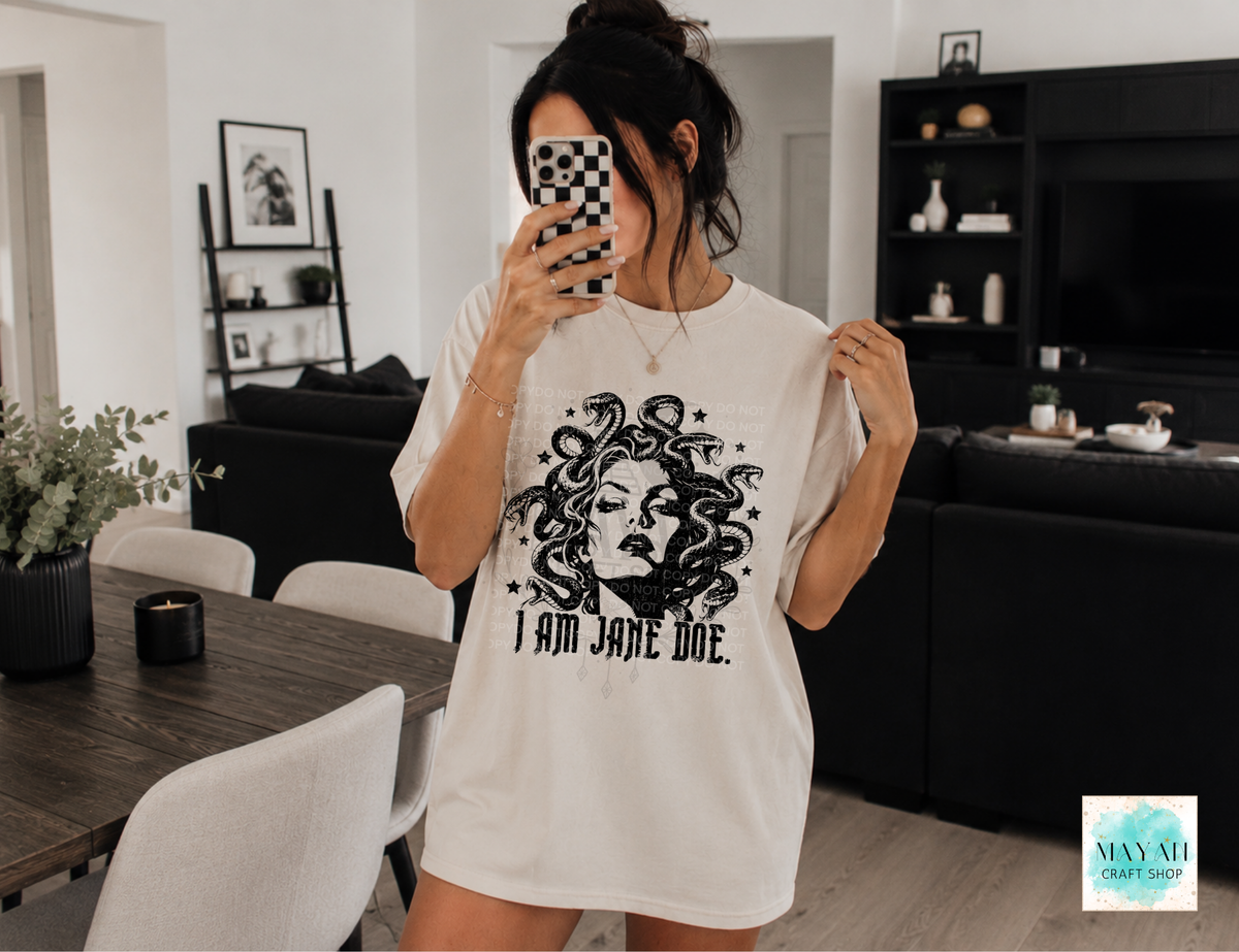 A woman takes a mirror selfie in a modern living room while wearing an oversized white graphic T-shirt with a black illustration of Medusa's face and the text 'I AM JANE DOE.' 