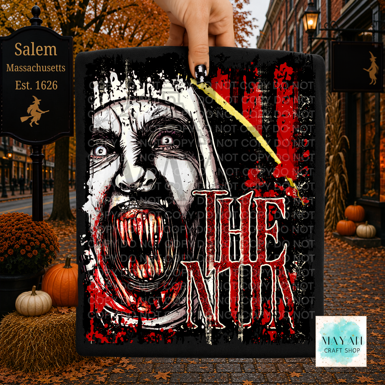 A hand holds a rectangular black Halloween sign featuring a screaming white-faced demon with red blood splatter. The sign is shown outdoors in an autumn street setting with pumpkins, fall leaves, and storefronts in the background.