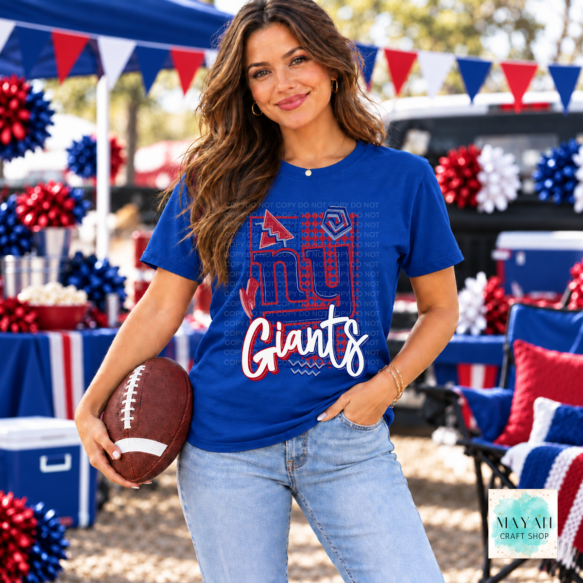 A woman wearing a royal blue football team graphic t-shirt and light wash jeans, holding a football in an outdoor patriotic tailgate setting. 