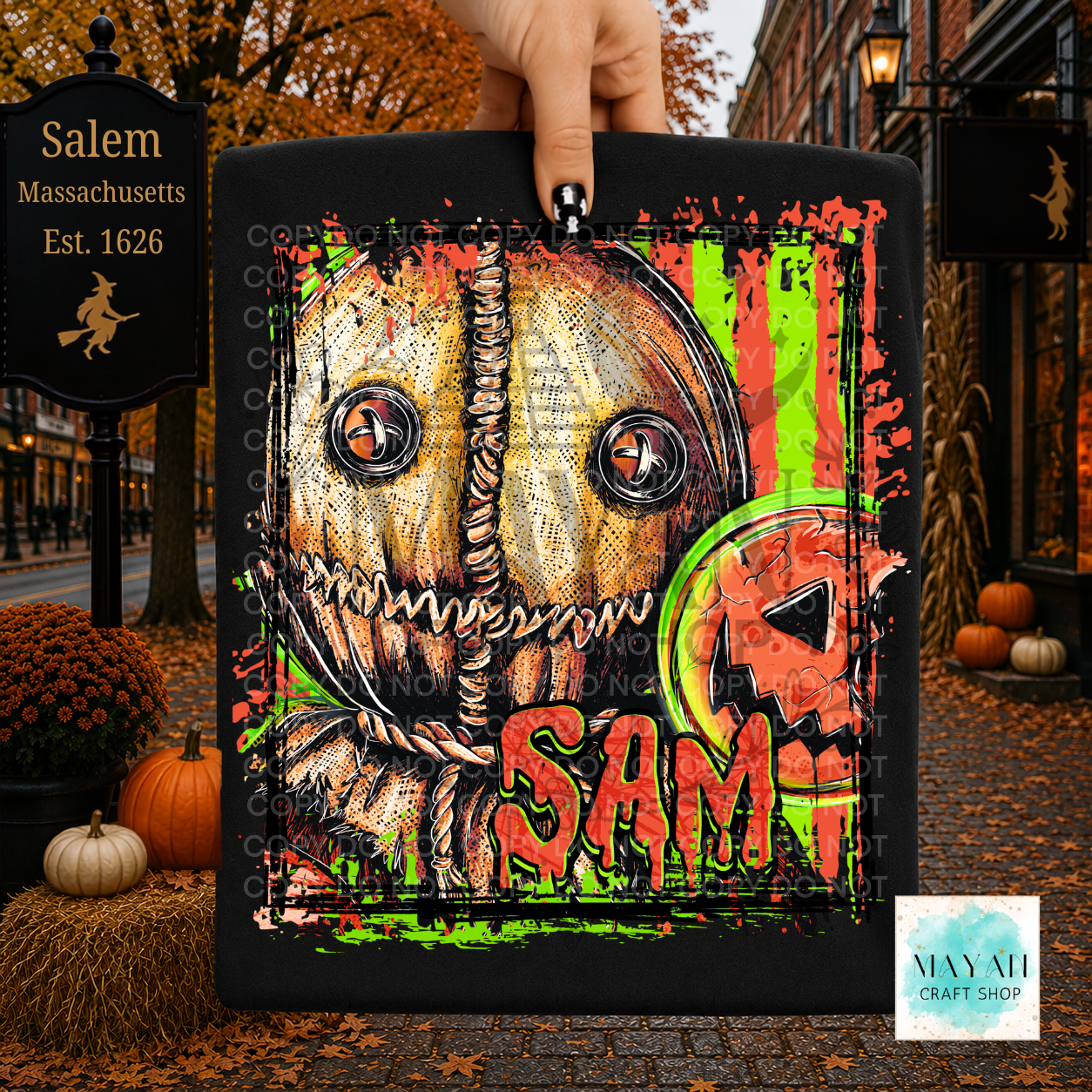 A hand holds a black shirt featuring a horror-style illustrated face with rope details, bright green vertical stripes, orange splatter accents. The background is a Halloween street scene with pumpkins, autumn leaves, storefronts, and a  sign.
