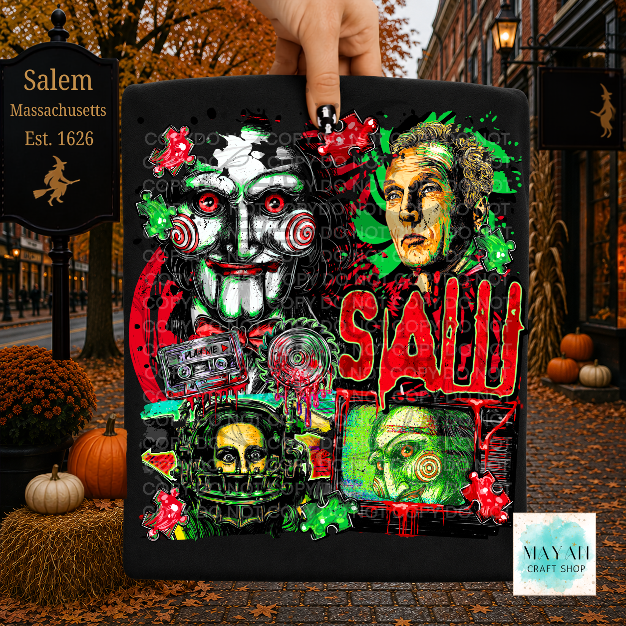 A hand holds a black square plaque featuring a colorful horror-themed collage, illustrated faces, skulls, and green/red paint splatter. The background is a Halloween street scene  with pumpkins, autumn leaves, and a signpost.