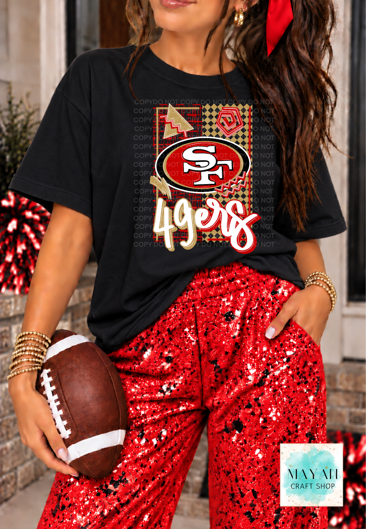 A woman wearing a black graphic T-shirt with a football team logo and red sequined pants, holding a football in a festive outdoor setting. 