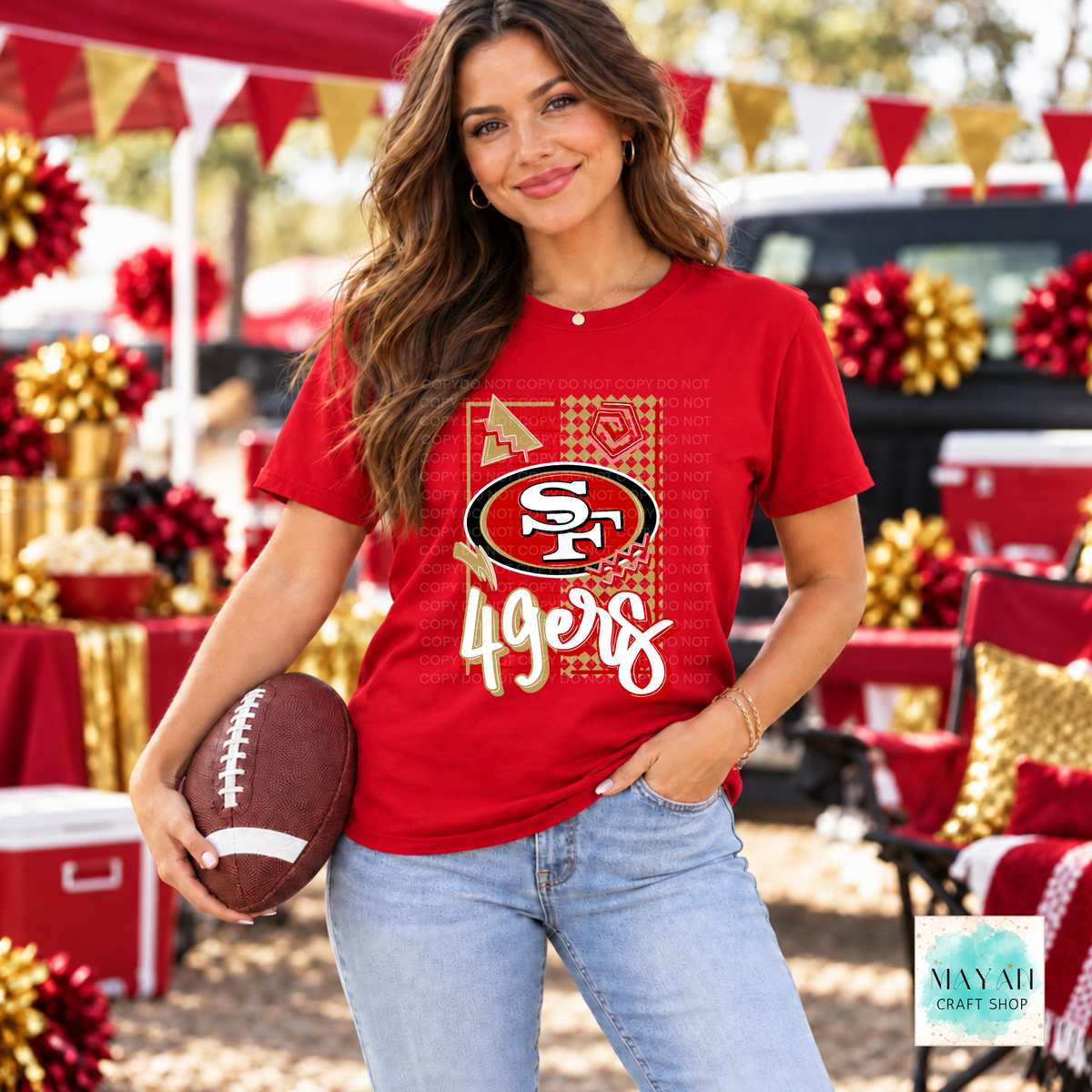 A woman wearing a red football team logo graphic t-shirt and light blue jeans, holding a football in an outdoor, festive, red, gold, and white color palette. The background shows a decorated tailgate scene with red and gold balloons, banners, and coolers.