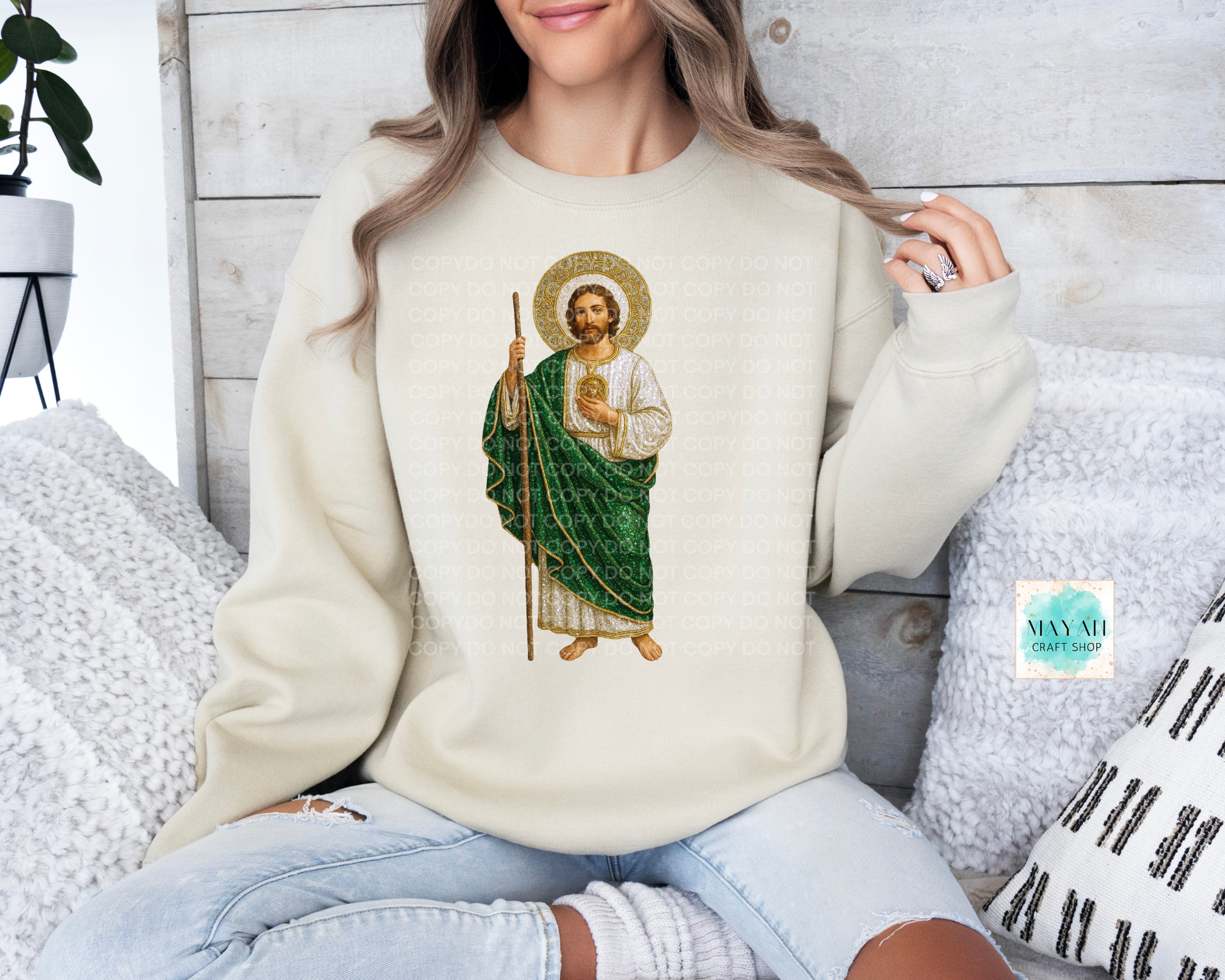 Person wearing a cream sweatshirt with a religious icon design, sitting on a textured surface.
