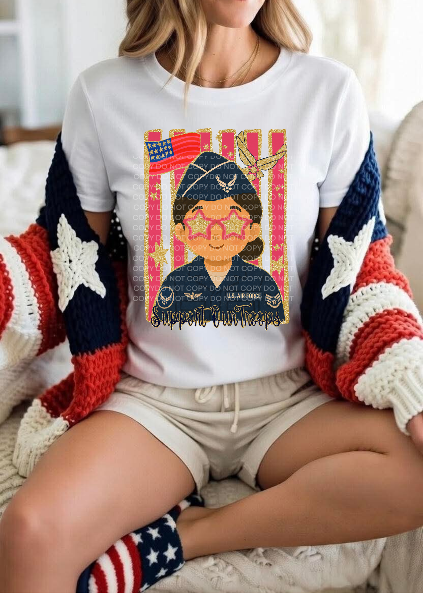 A woman sitting indoors wearing a white graphic T-shirt with a colorful military-themed print, a red, white, and blue knit cardigan, light shorts, and patriotic socks.