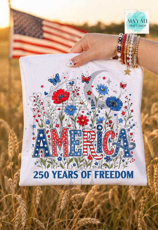 Hand holding a tote bag with 'America 250 Years of Freedom' design in a field with an American flag in the background.