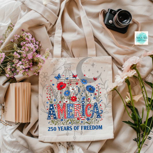 Tote bag with 'America 250 Years of Freedom' design on a fabric background with flowers and a camera.