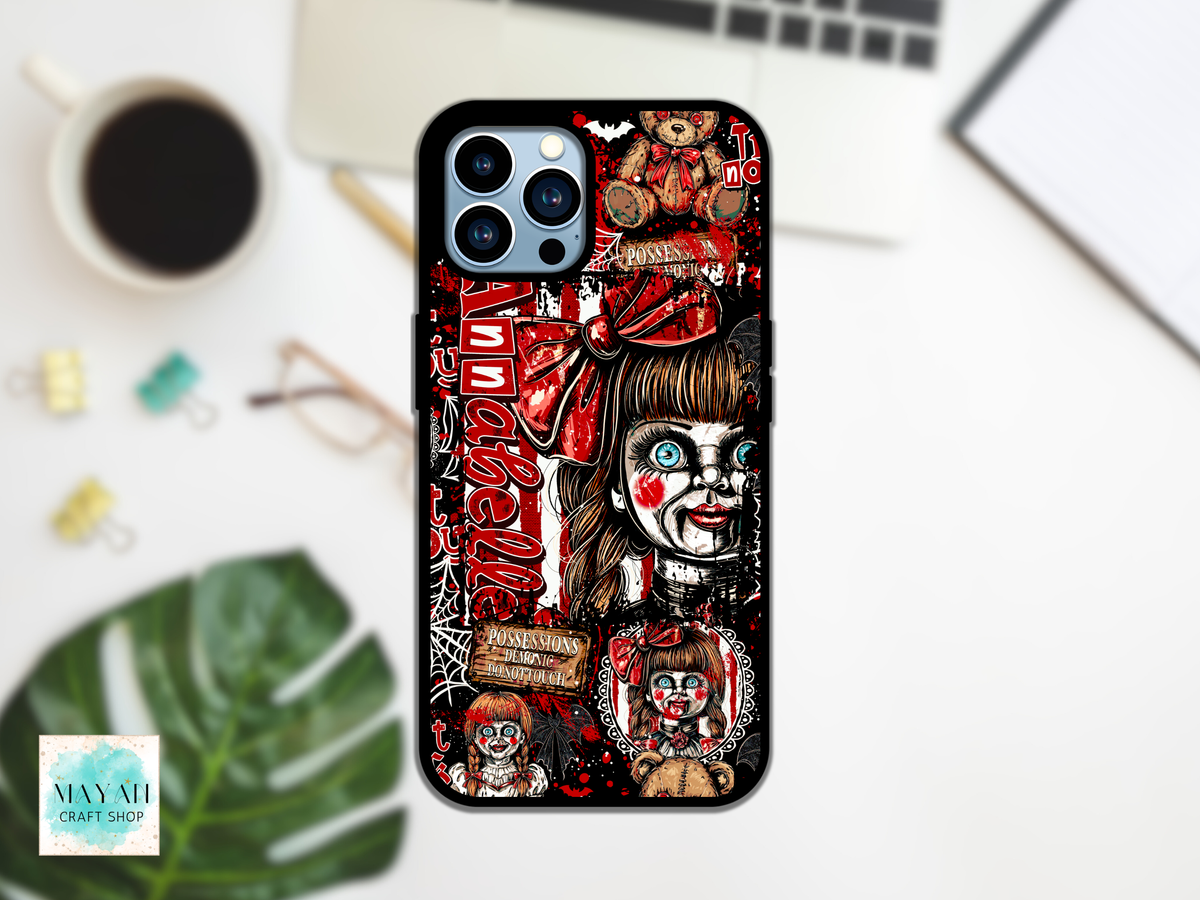 A black phone case featuring a detailed horror-themed collage design of a creepy doll with red makeup, a word in red script, and small teddy bear illustrations, shown on a white desk flat-lay with a coffee cup, glasses, and a laptop in the background.