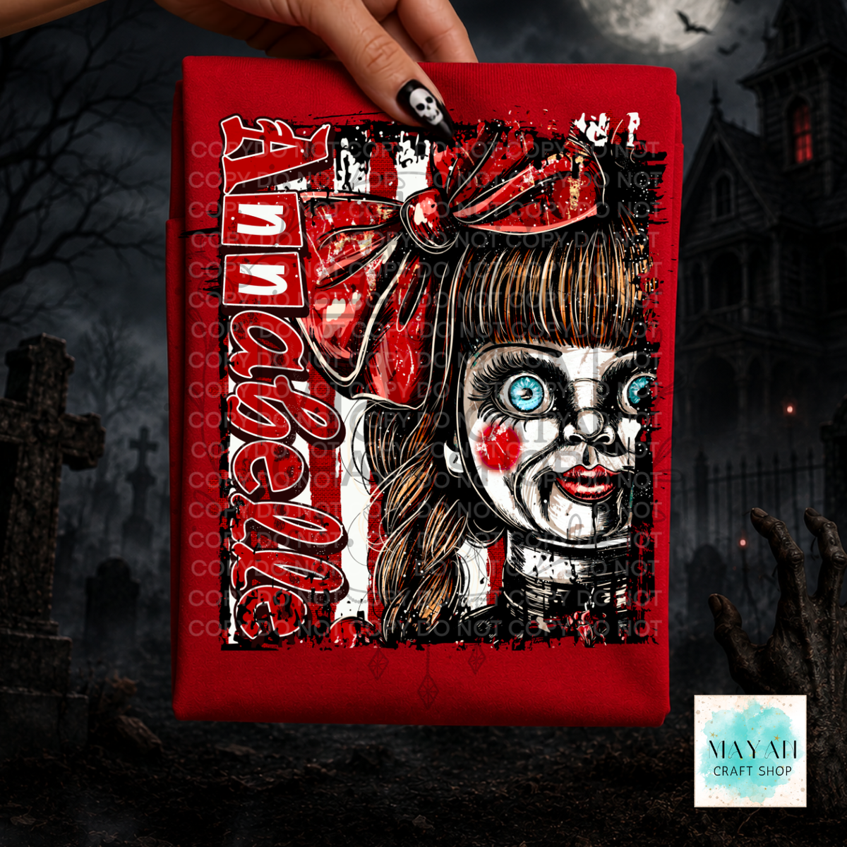 A hand holds a red shirt, likely a bandana or small flag, featuring a horror-style graphic of a doll-like face with blue eyes, a red bow, and a word in large red letters. The background is a dark, spooky graveyard scene with a haunted house.