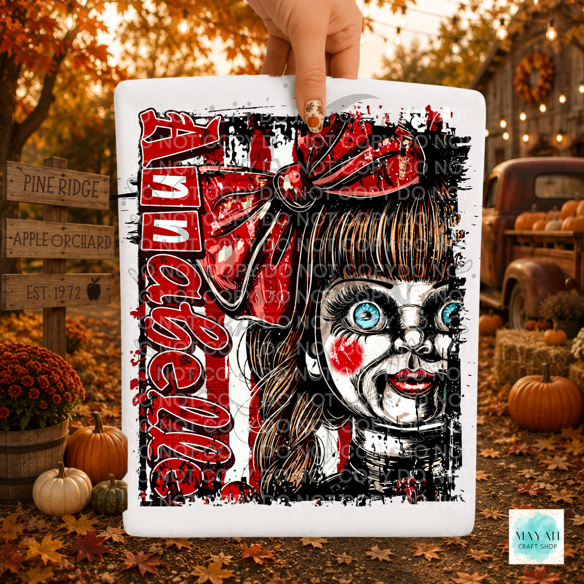 A hand holds a white rectangular sign featuring a stylized horror character with a red bow, blue eyes, and red vertical text on a white background. The scene is set outdoors in autumn with pumpkins, fall leaves, and rustic signage in the background.