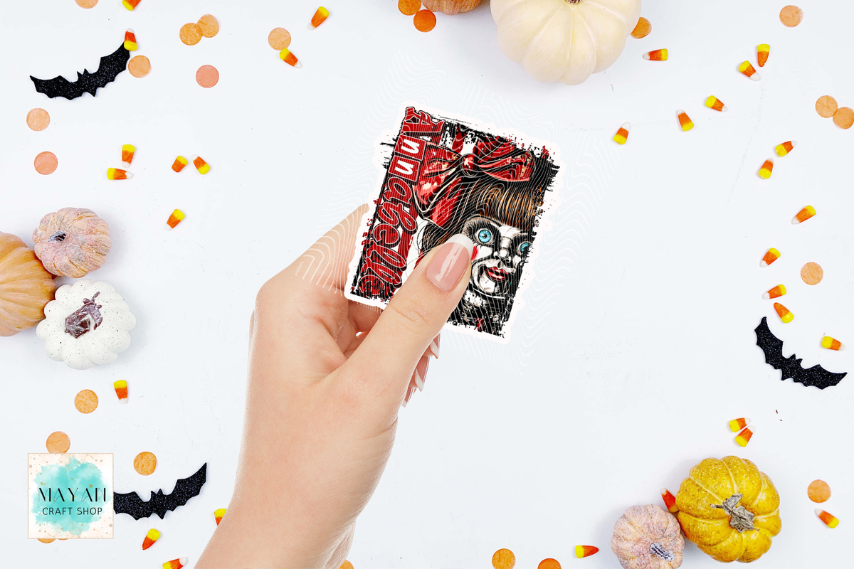 A hand holds a square art print featuring a stylized horror character with a word in red. The background is a white flat-lay styled with small pumpkins, candy corn, and black bat cutouts.