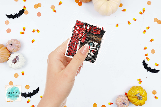 A hand holds a square art print featuring a stylized horror character with a word in red. The background is a white flat-lay styled with small pumpkins, candy corn, and black bat cutouts.